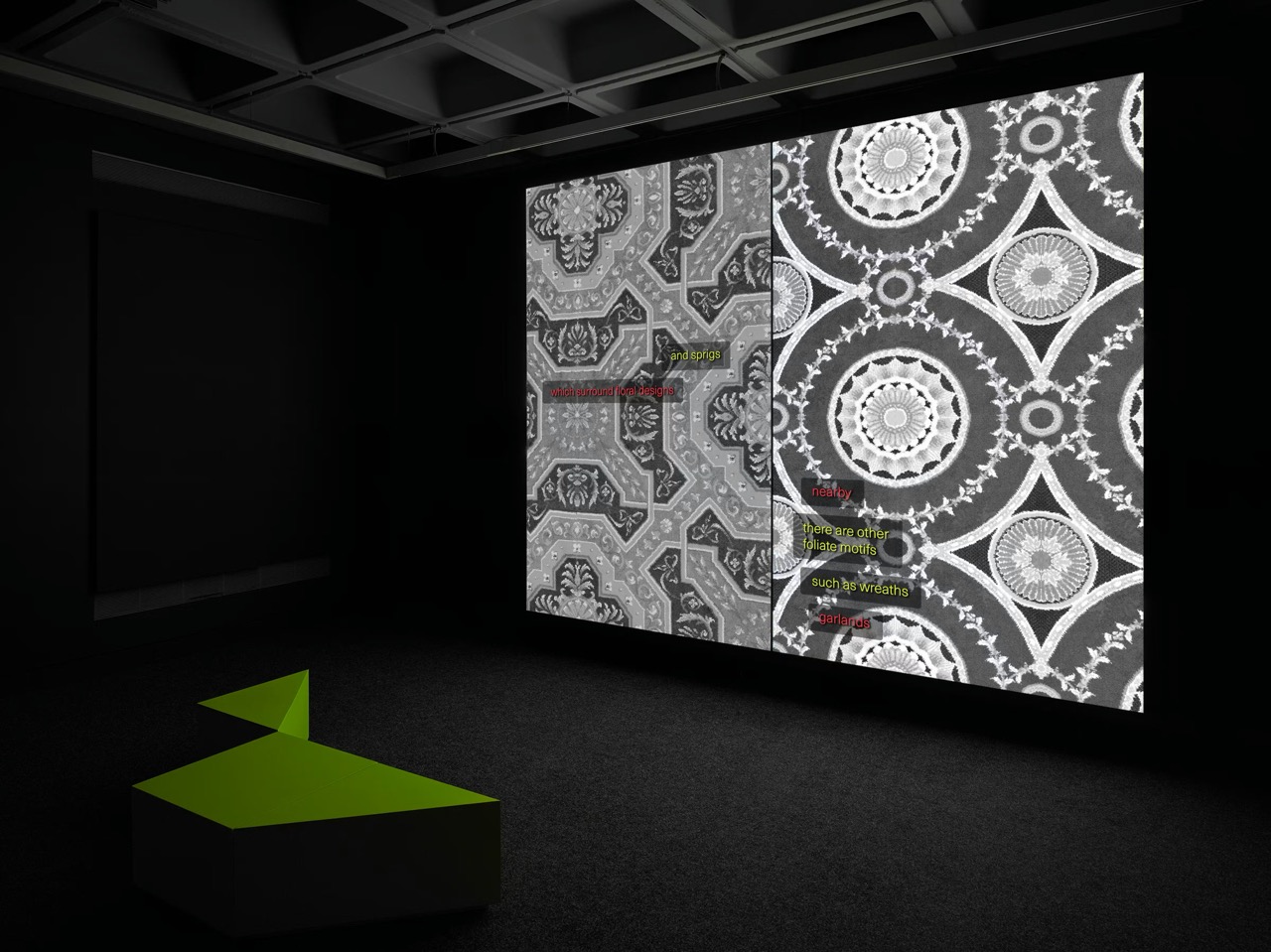 A dark gallery with a coffered ceiling. A large projection fills the wall with two ornate grey carpet patterns side by side, small lines of coloured text overlaid on them. An angular green bench sits on the dark carpet in front.