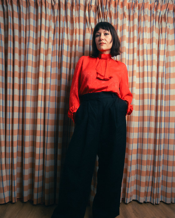 Leah Kardos in a red blouse and black trousers against a checked curtain, 2024