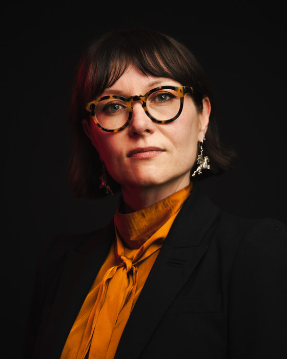 Leah Kardos in an orange tie-neck blouse and black blazer, lit from the side against black, 2026